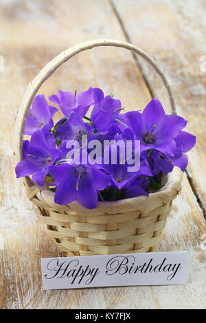 Happy birthday card with wicker basket filled with purple Campanula bell flowers on rustic wooden surface Stock Photo