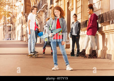 Portrait of smiling curly-haired teenage boy holding skateboard, spending holidays with friends at urban streets Stock Photo