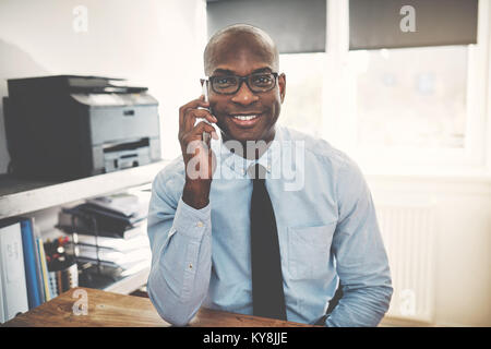 Smiling African entrepreneur sitting at a desk in his home office sending talking on a cellphone Stock Photo