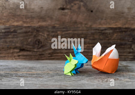 Three colorful origami Easter Bunny rabbits in green, blue, and orange made of paper, one stuffed with straw, cradling an egg for Easter or springtime Stock Photo