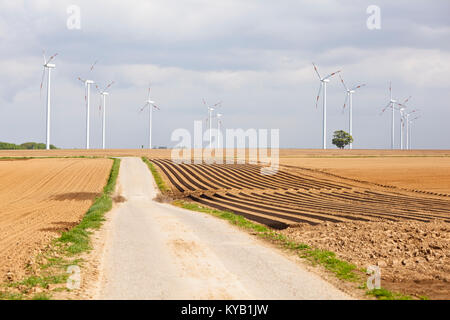 A row of wind turbines in agricultural landscape with a single tree and a road leading to them. Stock Photo