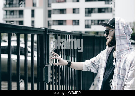 young man with beard in light checkered jacket with hood in a cap and sunglasses picks up the apartment number on the keypad to call the intercom Stock Photo