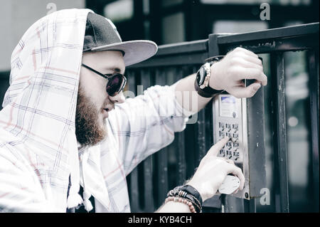 young man with beard in light checkered jacket with hood in a cap and sunglasses picks up the apartment number on the keypad to call the intercom Stock Photo