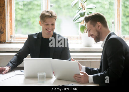 Two successful businessmen using laptops talking discussing at meeting, young entrepreneurs in suits negotiate sitting at office desk with computers, Stock Photo