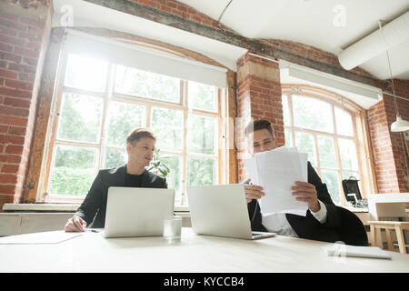 Business partners discussing papers sitting at table meeting in big loft office, two serious businessmen wearing suits analyzing project statistics gr Stock Photo