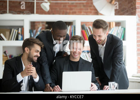 Multi-ethnic smiling businessmen in suits watching something funny on laptop in office together, diverse excited business team happy to see good news Stock Photo