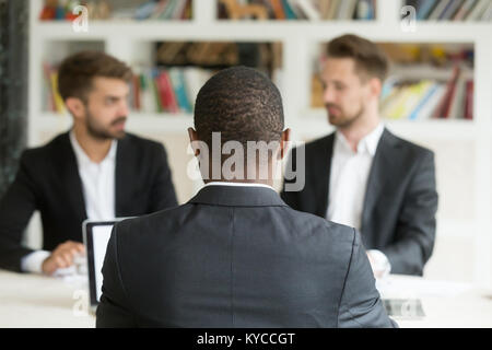 Rear view at african businessman against two caucasian men wearing suits, black vacancy candidate sitting his back to camera on job interview with two Stock Photo
