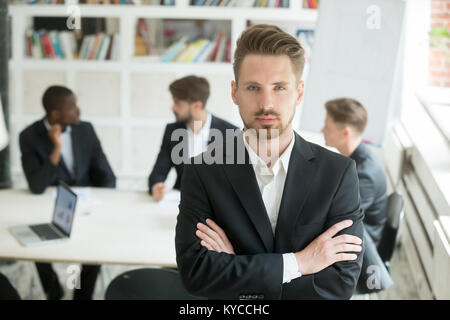 Serious young leader in suit looking at camera standing arms crossed on executive team meeting background, confident manager boss ceo co owner posing Stock Photo