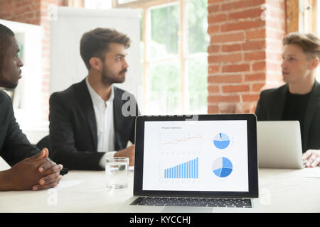 Project statistics on laptop screen with businessmen talk negotiate at background, executive team discussing project development work result, analyzin Stock Photo