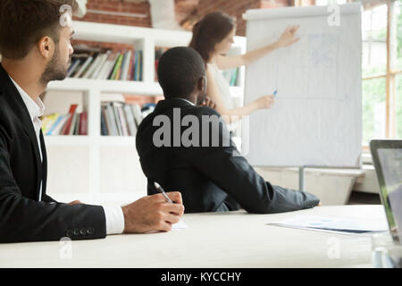 Diverse focused businessmen in suits listening to female coach giving presentation on flipchart sitting making notes at conference meeting table, corp Stock Photo