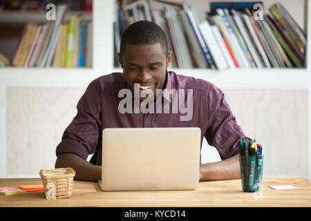 Smiling african-american man using laptop sitting at home office desk, happy excited black e business owner looking at computer screen pleased by read Stock Photo