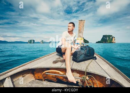 Young man (tourist) with backpack on the traditional long tail boat against islands between Phuket and Krabi in Thailand. Stock Photo