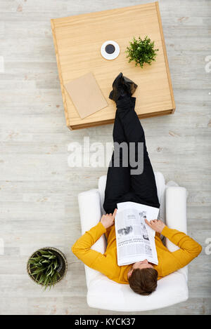 Directly above view of dark-haired man wearing mustard sweater sitting on cozy armchair and taking nap, newspaper lying on his belly Stock Photo