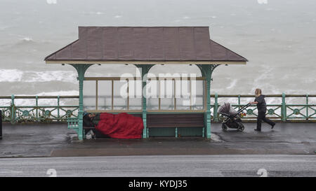 Brighton, UK. 15th Jan, 2018. A homeless man shelters on Brighton seafront as a woman with a baby walks by as gales and heavy rain batter the south coast this morning with colder weather forecast to hit Britain later in the week Credit: Simon Dack/Alamy Live News Stock Photo