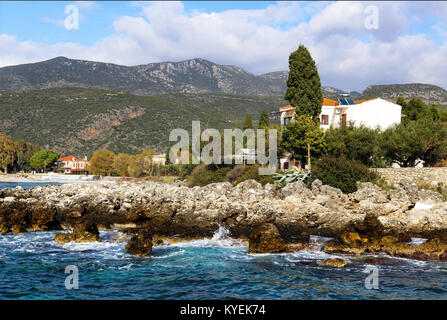The very blue Ionian Sea splashes on the rocks of the Peloponnese Peninsula in Greece with tall moutains towering behind the red tiled houses near the Stock Photo