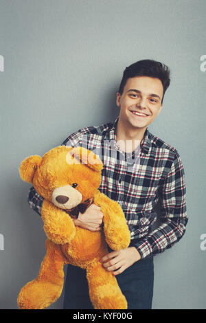 Man holds teddy bear on a light blue background, copy space. Pretty toy concept. Man hugs cute toy bear tight. Guy with happy face plays with red soft toy. Vintage filter Stock Photo