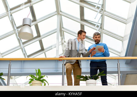 Portrait of two successful business men standing on balcony in modern office building and using digital tablet for work  under glass roof, copy space Stock Photo