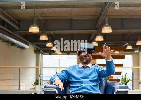 Portrait of mature bearded man wearing VR headset sitting in armchair looking amazed and excited reaching out with his hands, copy space Stock Photo