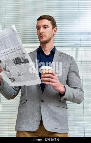 Waist up portrait of handsome young businessman reading newspaper and drinking coffee standing against glass wall in office Stock Photo