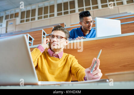 Low angle view of two students, one of them Middle-eastern, sitting at desks in modern auditorium at college and preparing for class, focus on young m Stock Photo