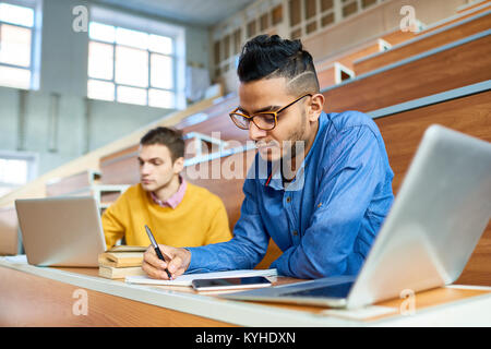 Portrait of two students sitting at desks in modern auditorium at college and preparing for class, focus on young Middle-Eastern man writing in copybo Stock Photo