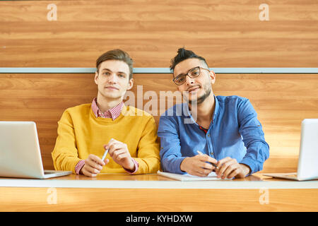 Portrait of two students sitting at desk in modern auditorium at college and looking at camera, one of them Middle-Eastern Stock Photo