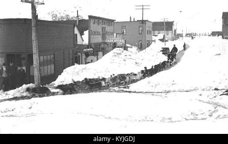 The photograph shows a dog sled transporting passengers through snow-covered streets in Valdez, Alaska, around 1912, illustrating winter transportation methods and local life. Stock Photo