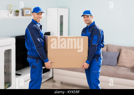 Two Young Smiling Movers In Uniform Delivering Cardboard Boxes In Living Room Stock Photo