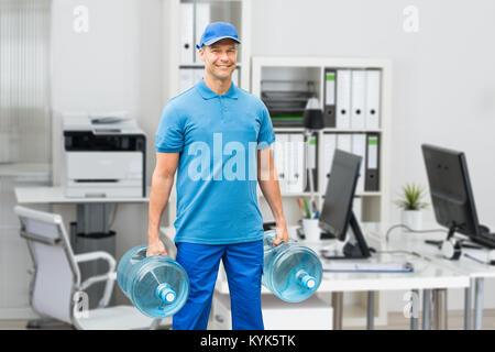 Portrait Of A Smiling Mature Delivery Man Holding Large Water Bottle In Office Stock Photo