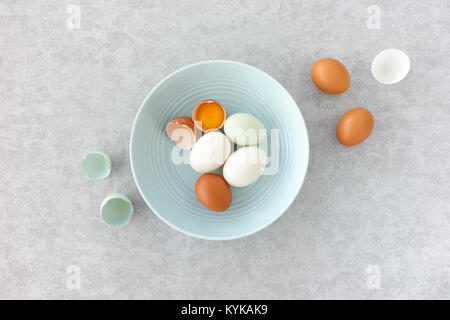 Blue eggs, brown eggs and duck eggs, whole and halves, in a pastel blue bowl on grey textured background. Top view. Stock Photo