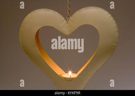Burning tealight candle in a heart shaped metal candle holder hanging in front of a graduated plain background Stock Photo