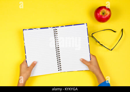 Kid hand with open notebook, glasses and red apple on yellow background. Stock Photo