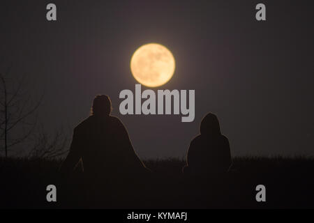 Two women are sitting on the ground against the horizon, the full moon rising between them. Stock Photo