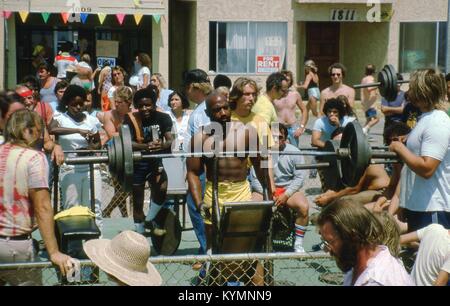 Scene of people exercising at the Muscle Beach Gym outdoor weight pen, located on Ocean Front Walk in Venice, California, June, 1978. At center is an African American man preparing to lift a large barbell. Crowds of onlookers watch the workout from both sides of a chain-link fence along the boardwalk. Visible in the background are storefronts at 1809 and 1811 Ocean Front Walk. () Stock Photo