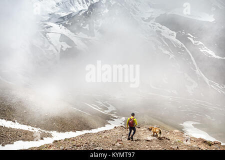 Hiker in green shirt with backpack and dog walking in the snowy mountains at foggy sky background Stock Photo