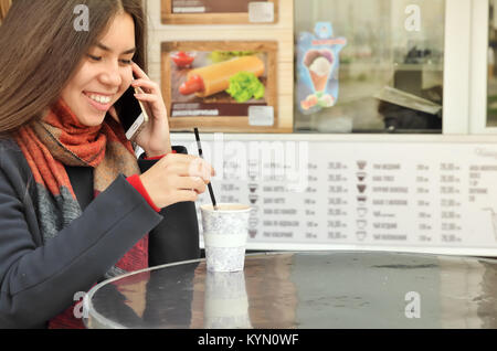 An attractive Asian girl drinks coffee and speaks by telephone at the coffee shop at the table. Dressed in a coat and a scarf Stock Photo