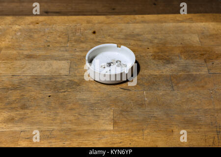 Used dirty white ceramic cigarette ash tray on a slated wooden table top. Stock Photo
