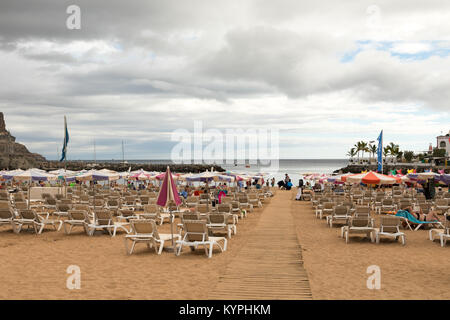 Puerto de Mogan, Gran Canaria in Spain - December 16, 2017: Sunbeds and parasols on the beach, with a board walk in the sand. Cloudy sky. Stock Photo