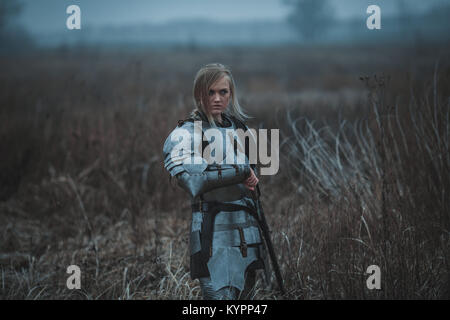 Girl in image of Jeanne d'Arc in armor and with sword in her hands stands on meadow in middle of dry grass. Stock Photo