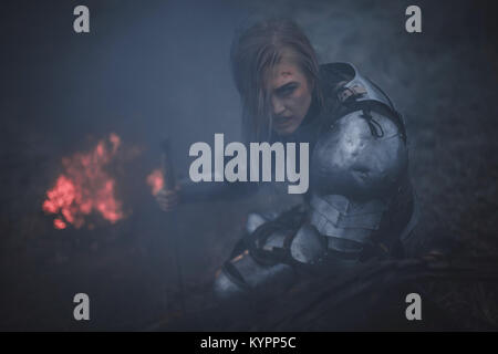 Girl in image of Jeanne d'Arc in armor and with sword in her hands kneels against background of fire and black smoke. Closeup. Stock Photo