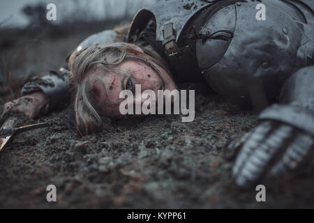 Girl with wounds on her face in image of Jeanne d'Arc in armor lies in mud with sword in her hands. Stock Photo