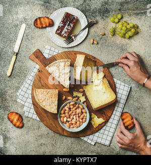 Flat-lay of cheese platter with cheese assortment, green grapes, honey and nuts with female hands reaching to food over grey concrete background, top Stock Photo