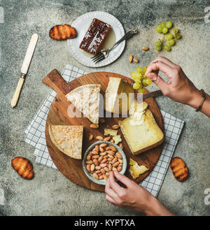 Flat-lay of cheese platter with cheese assortment, green grapes, bread, honey and nuts with female hands reaching to food over grey concrete backgroun Stock Photo