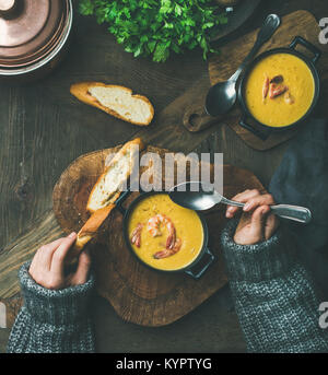 Woman in sweater eating corn creamy soup with shrimps in individual pot, top view. Woman' s hand keeping spoon and bread slice. Flat-lay of rustic din Stock Photo