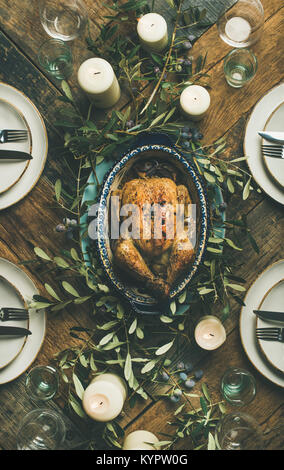 Flat-lay of whole roasted chicken in tray for Christmas eve celebration, plates, glasses and candles over rustic wooden background, top view, vertical Stock Photo