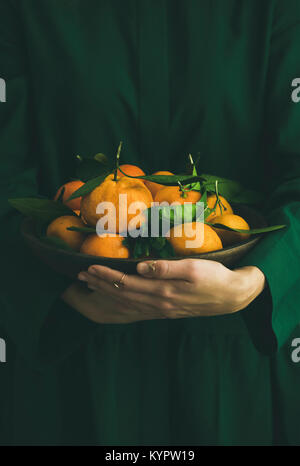 Fresh raw tangerines citrus fruits with leaves in plate in hands of girl wearing green dress, selective focus, copy space. Winter Christmas of New yea Stock Photo