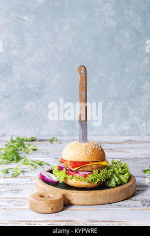 Homemade burger in classic bun with tomato sauce, arugula, meat, cheese, onion on wood serving board over white wooden plank table. Rustic style. Home Stock Photo
