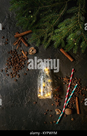 Christmas lights garland in glass bottle with coffee beans, spices, nuts, cocktail tube, fir tree branches in wooden tray over dark texture background Stock Photo