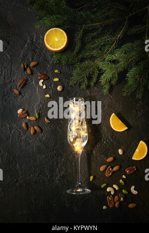 Christmas lights garland in champagne glass with nuts, orange, fir tree branches over dark texture background. Christmas holiday mood card. Top view w Stock Photo