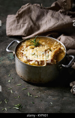 Traditional french onion soup with cheese and bread served in vintage aluminum pan with rosemary and textile napkin over dark metal background. Stock Photo